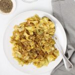 White plate with cooked cabbage and onions with spoon and gray napkin.