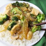 A white plate with rice and tofu veggie stir fry with sesame seeds, a green napkin, and chopsticks.